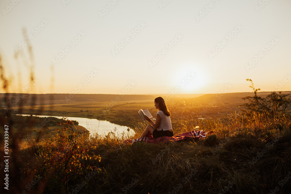 Calm girl reading a book on hill with perfect landscape, enjoying time on holiday. She read book in quiet nature.