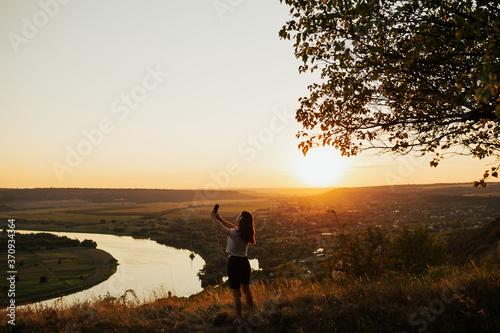 Woman on amazing summer sunset. Girl taking a selfie on a mountain viewpoint. Get the power of nature at the sunset at the mountain. 