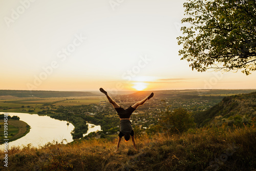 Man doing handstand on the grass at sunset sky. Young sporty man doing handstand exercise in beautiful mountain landscape. Sports outdoor lifestyle. Copy space.