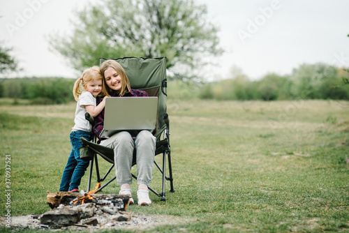 Mother work on Internet with kid outdoors. Quarantine, closed nursery school during coronavirus outbreak. Communication with family online on laptop near fire in nature. Homeschooling, freelance job.