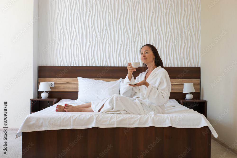 Relaxed middle aged woman with brown hair, wearing white bathrobe. The woman sits on her bed drinking tea or coffee from the white mug, rest in hotel concept.