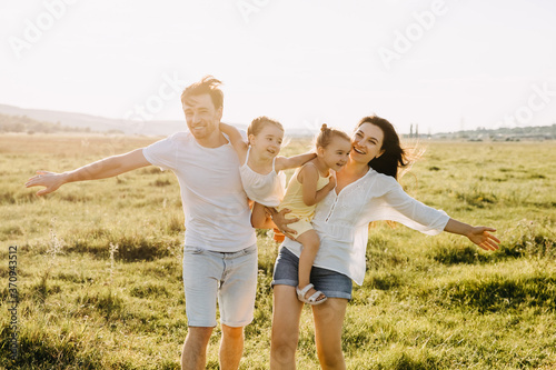 Family of four playing in a field on a summer day. Mother, father and two daughters running on green grass, pretending to be airplanes.