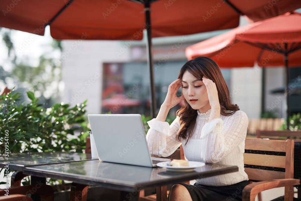 Adult Asian woman is working on a laptop in a cafe outside on the street having a headache feeling sick holding her hands on her head Adult Asian woman is working on a laptop in a cafe outside on the street having a headache feeling sick holding her hands on her head