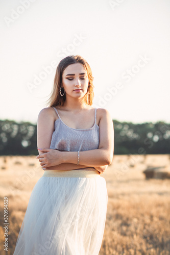young woman in dress walking in evening in field with hay bales, beautiful romantic girl with long hair outdoors in field