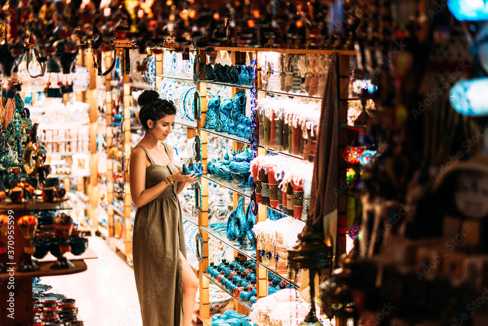 Fototapeta premium Beautiful girl in a souvenir shop in Turkey. Girl chooses a souvenir Oriental shop. Turkish Souvenirs. Oriental bazaar. Turkish Bazaar. Gift shop in Goreme, Turkey. Moroccan Bazaar. Copy space.