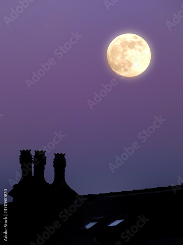 a glowing full moon in a purple twilight summer sky above a house rooftop in silhouette