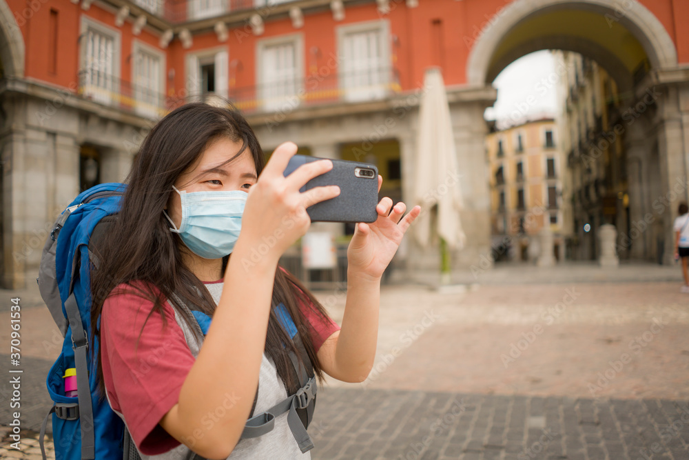 new normal Asian backpacker girl enjoying holidays travel in Europe - young happy and beautiful Chinese tourist woman in mask taking photo with mobile phone in city tour