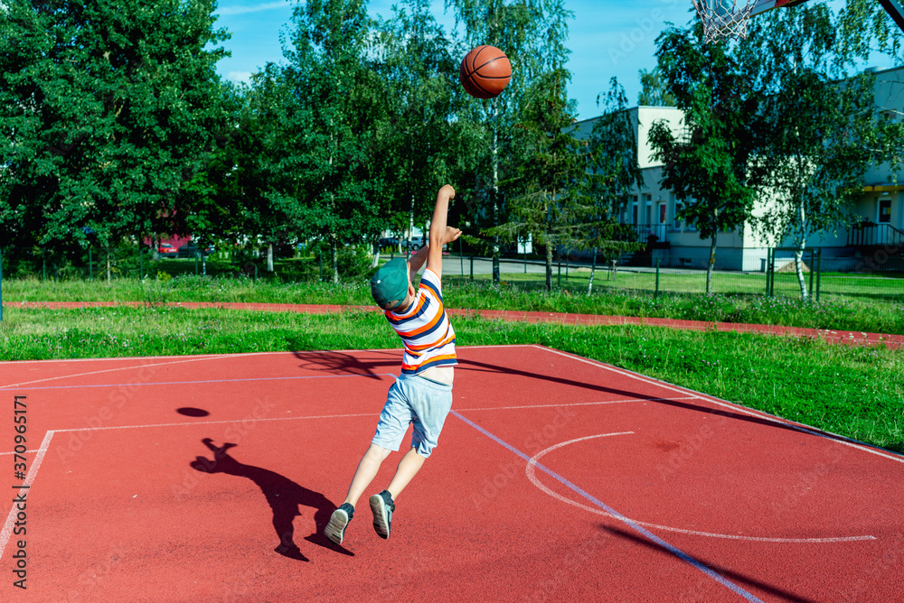 Attractive east european caucasian little basketball player jumps to throw the ball into the hoop.outdoors red court