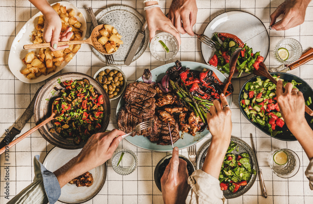 Summer barbeque party. Flat-lay of table with grilled meat, vegetables, salad, roasted potato and peoples hands with cutlery over white tablecloth, top view. Family gathering, comfort food concept - obrazy, fototapety, plakaty 