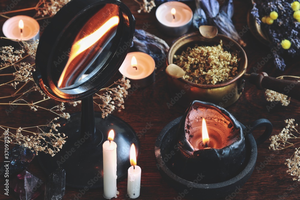 Candle flame divination in concave mirror on wiccan witch altar. Black candle burning on a messy cluttered table with dried herbs and flowers, crystals, plants, other nature objects in dark background - obrazy, fototapety, plakaty 