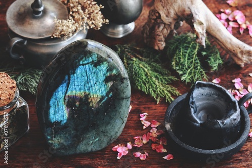 Labradorite crystal on dark wooden table with various nature objects like dried evergreens, plants, flowers, herbs in the background, Messy and cluttered wiccan witch altar with big labradorite on it photo