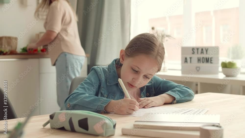 Medium shot of blond-haired Caucasian schoolgirl sitting at kitchen table and doing homework while her mother cooking on background. Then woman coming and starting helping her daughter
