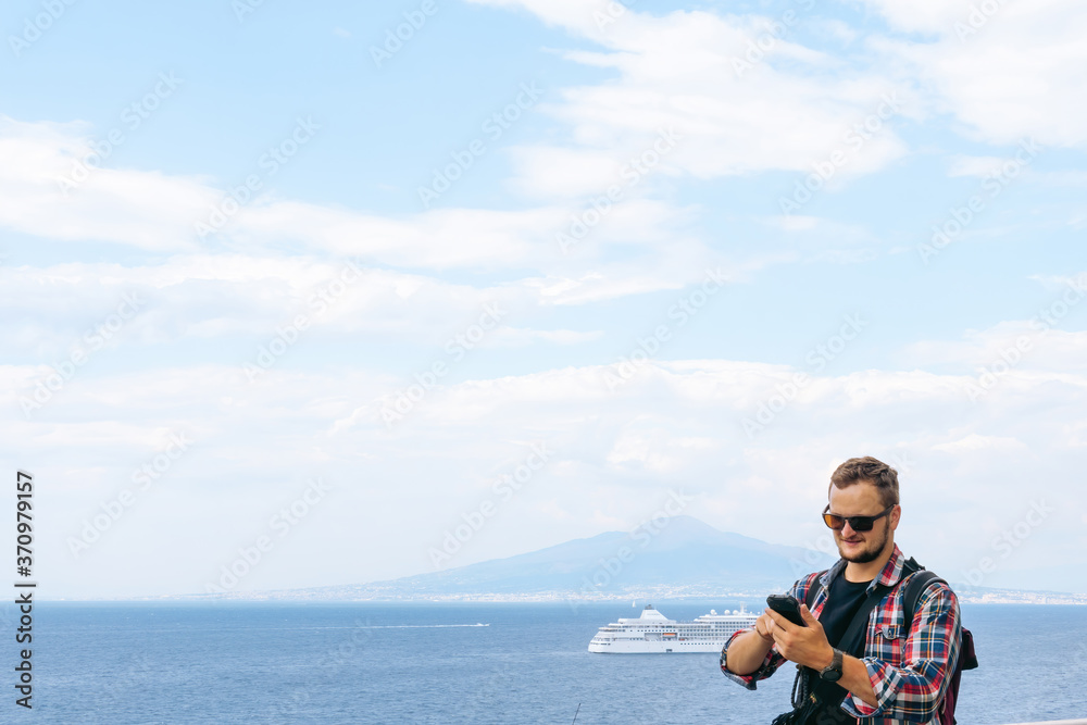 Male tourist with glasses and a banana bag, uses a smartphone. Communication and mobile internet concept. Roaming. View of the sea and Mount Vesuvius. Ocean liner.