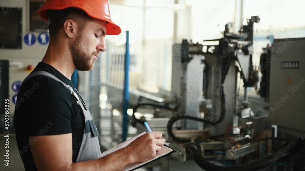 Manager in orange hard hat stands in the factory with machines and writes information in notepad. Manager in orange hard hat stands in the factory with machines and writes information in notepad.