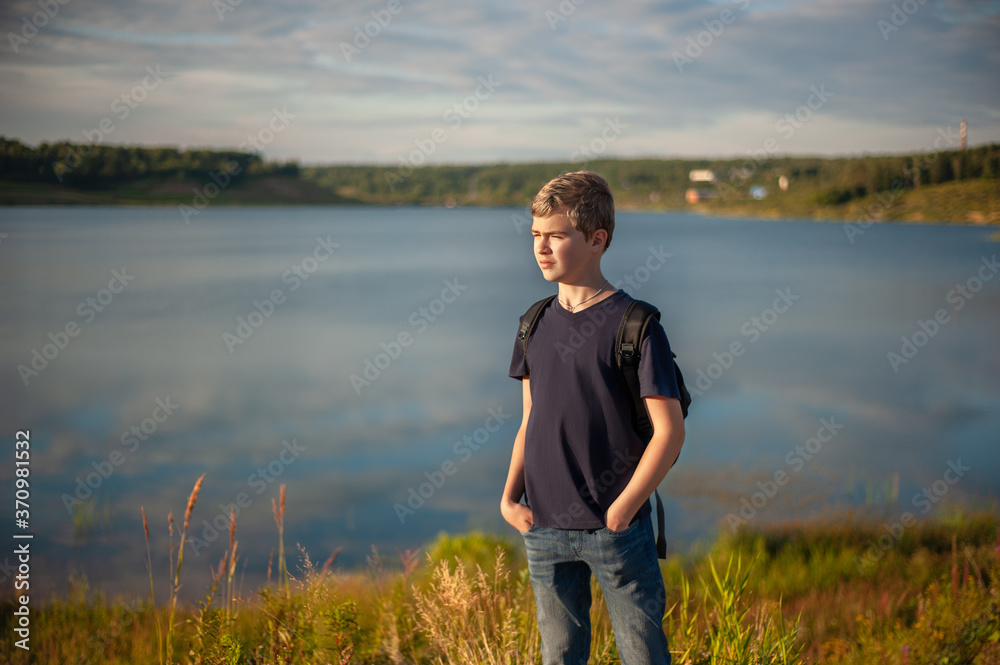 A teenage boy with a backpack on the shore of a beautiful lake in the evening at sunset.