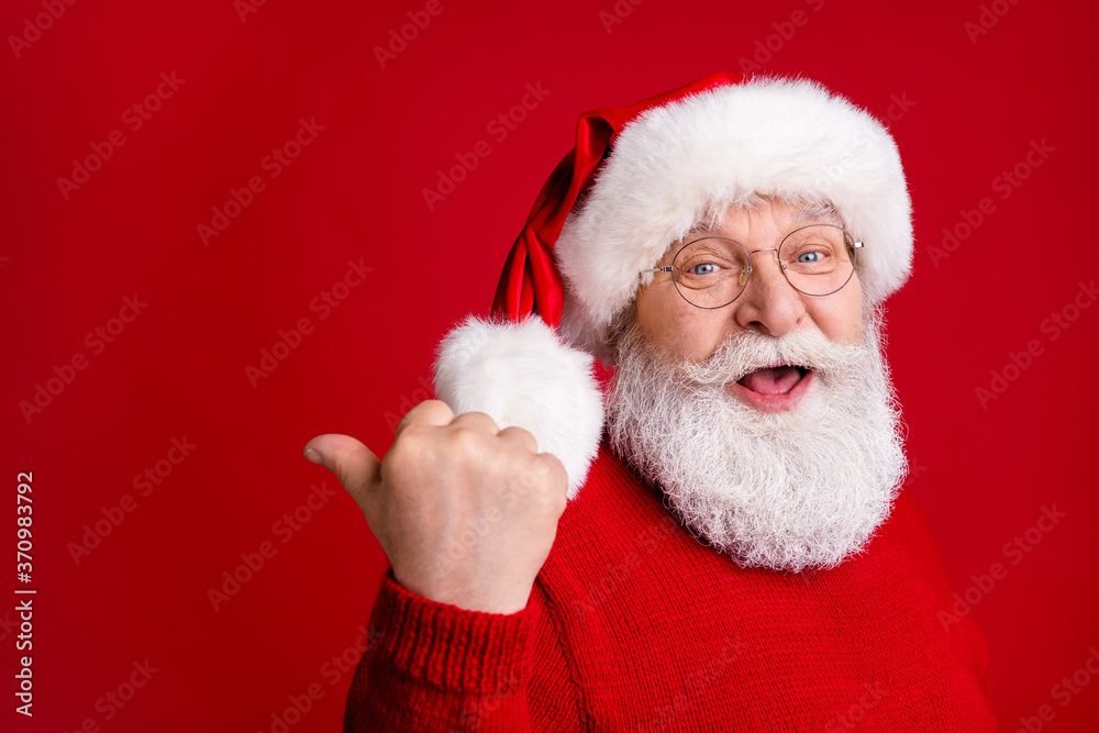 Photo of amazed old man in santa claus headwear point thumb finger copyspace demonstrate holly jolly miracle discounts wear x-mas sweater jumper isolated bright shine color background