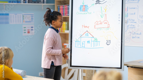 Elementary School Science Class: Portrait of Cute Girl Uses Interactive Digital Whiteboard to Show to a Full Classroom how Renewable Energy Works. Science Class, Curious Kids Listening.