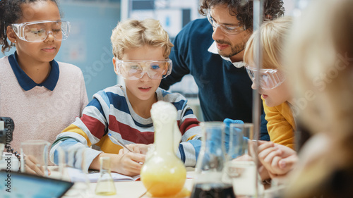 Elementary School Science Classroom: Enthusiastic Teacher Explains Chemistry to Diverse Group of Children, Little Boy Mixes Chemicals in Beakers, Writes down Results. Children Learn with Interest