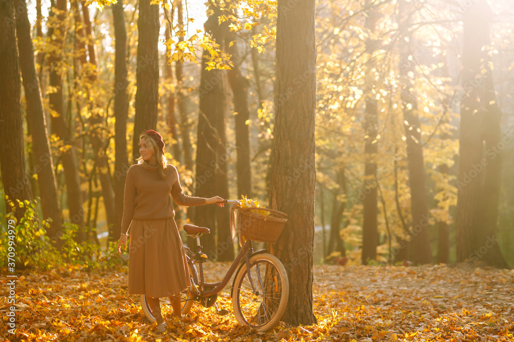 Stylish woman  with a bicycle enjoying autumn weather in the park. Beautiful Woman walking  in the autumn forest at sunset. - obrazy, fototapety, plakaty 