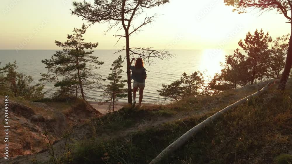 Young woman enjoys beautiful scenic sunset at Baltic Sea standing at tree on hill. Slow aerial orbit Young woman enjoys beautiful scenic sunset at Baltic Sea standing at tree on hill. Slow aerial orbit
