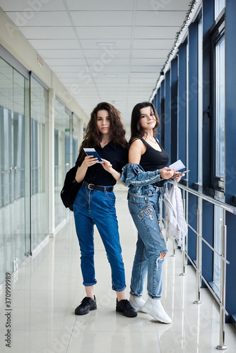 Two young brunette girls, standing in light airport hallway with huge windows, wearing casual jeans clothes, holding international passports and boarding passes tickets. Girlfriends traveling by air.