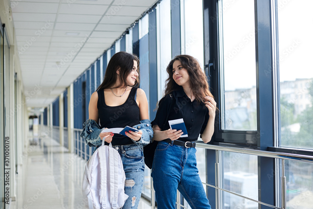 Two young brunette girls, standing in light airport hallway with huge windows, wearing casual jeans clothes, holding international passports and boarding passes tickets. Girlfriends traveling by air.