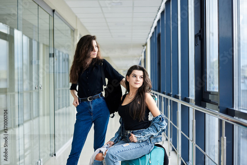 Two young brunette girls  sitting on small mint carry on luggage in light airport hallway with huge windows  wearing casual jeans clothes. Girlfriends  traveling by air  waiting for flight.