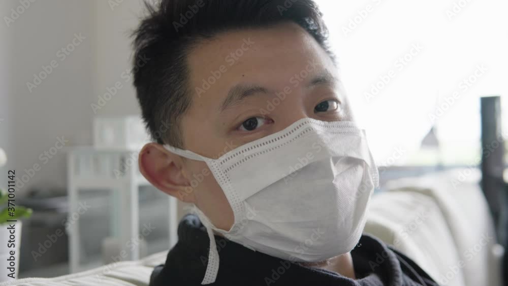 A young Asian man in a face mask turns and looks at the camera as he sits on a couch at home - face closeup A young Asian man in a face mask turns and looks at the camera as he sits on a couch at home - face closeup