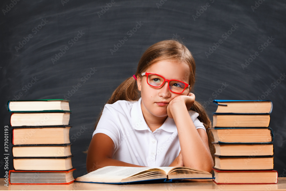 Back to school. Little girl student sitting at the table on the background of a black board. Tired schoolgirl with piles of books