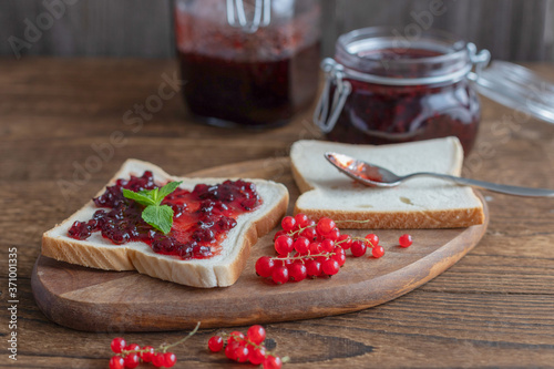 Homemade red currant jam in a glass jar and a number of berries on a wooden background. Toast or bread with marmalade for breakfast. Preparations for the winter from berries and fruits