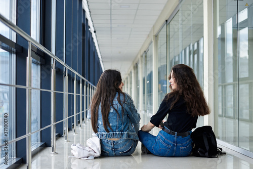 Two young brunette girls, sitting on floor in light airport hallway, with backs to camera, wearing casual jeans clothes. Girlfriends, traveling by air, waiting for flight.