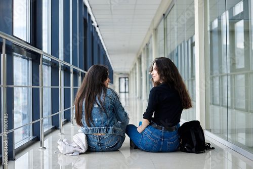 Two young brunette girls, sitting on floor in light airport hallway, with backs to camera, wearing casual jeans clothes. Girlfriends, traveling by air, waiting for flight.
