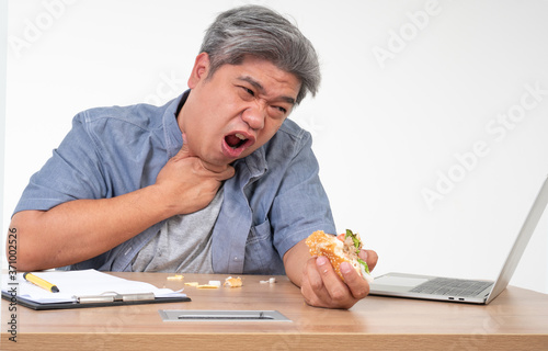 Bild auf Leinwand Asian man working and eating a burger on office desk and holding his neck after choking foods