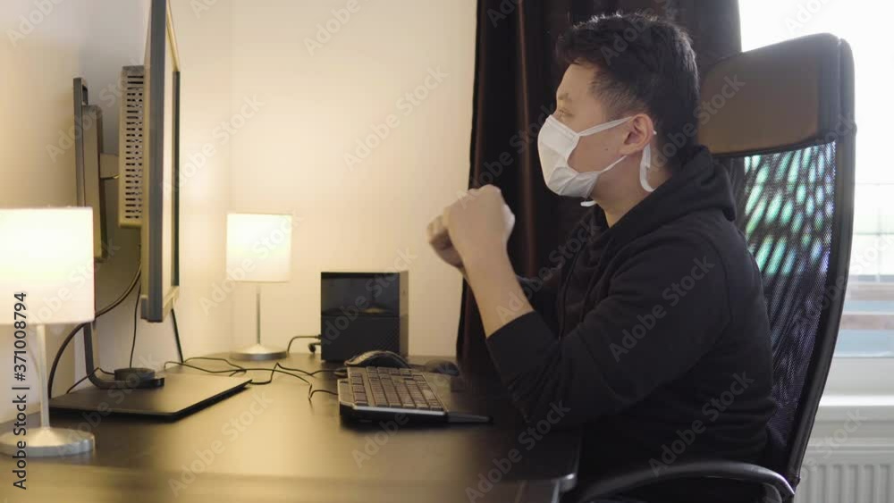 A young Asian man in a face mask celebrates as he looks at a desktop computer screen at home A young Asian man in a face mask celebrates as he looks at a desktop computer screen at home