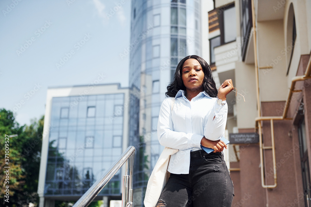 With phone in hands. Young afro american woman in white shirt outdoors in the city against business building With phone in hands. Young afro american woman in white shirt outdoors in the city against business building