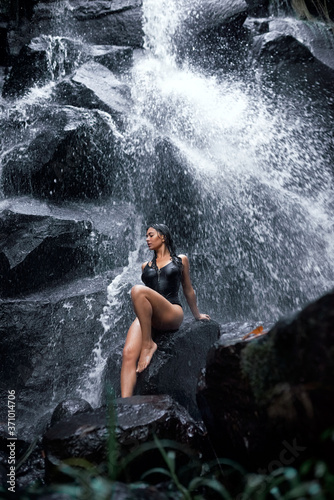 beautiful brunette sits against the background of a waterfall and washes under it.