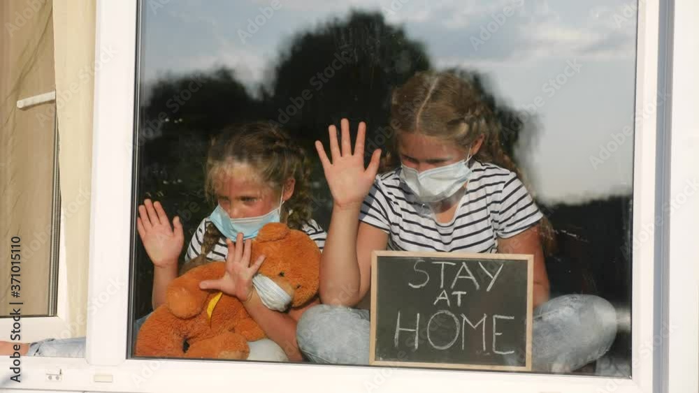 stay at home coronavirus. kids in medical mask in home with a toy teddy bear. kids holding a sign with the inscription stay at home. quarantine sitting by the window. coronavirus concept covid 19
