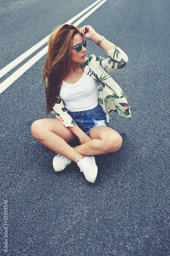 Beautiful Californian hipster girl sitting on her cruiser longboard in the middle of asphalt road, trendy fashion model posing with skate board wearing colorful sunglasses, copy space area for content