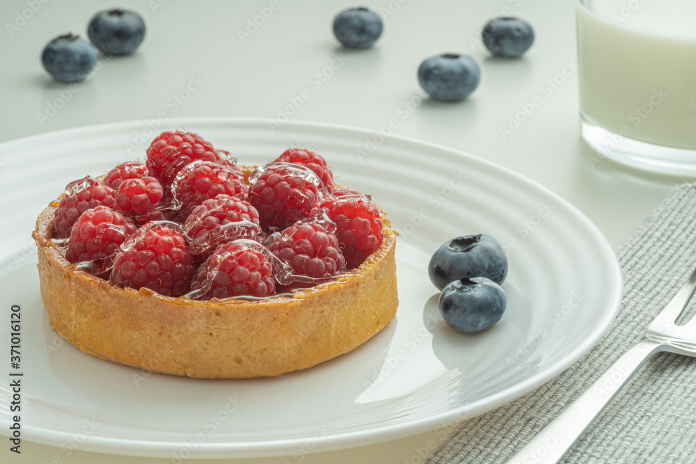 Raspberry tartlets served on a transparent plate with brown sugar. Glass of milk in the sun on a white background. Raspberry tartlets served on a transparent plate with brown sugar. Glass of milk in the sun on a white background.