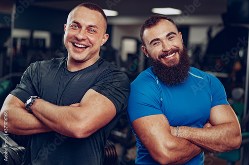 Two smiling fit men bodybuilders standing side to side in a gym