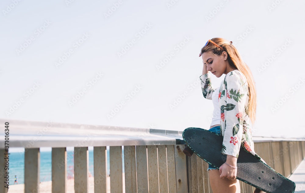 Stylish female skater holding her longboard while standing on pier near the beach against blue sky background with copy space area for your text message or content, hipster girl relaxing after riding