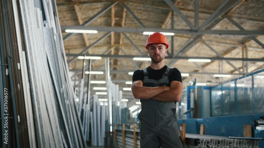 Worker in grey uniform and hard hat stands indoors in the factory with arms crossed.