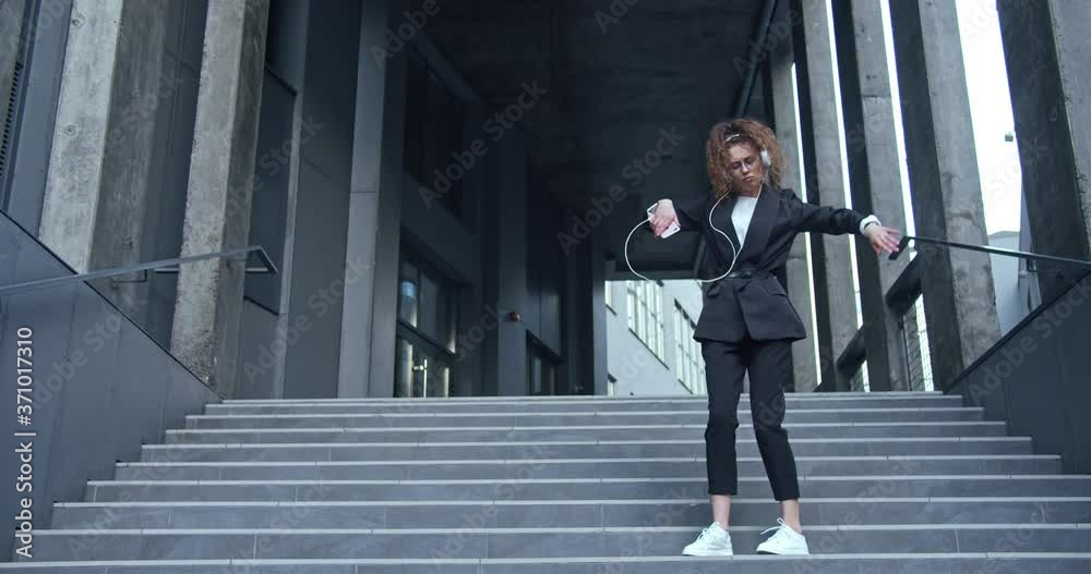 Attractive joyful business lady wearing a formal suit in headphones and glasses. Charming young office female worker havimg fun, enjoying, listening to music and actively dancing outdoors.