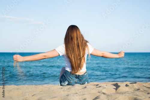 Portrait of a young hipster girl with luxurious long hair sitting with open arms in front of beautiful calm sea and blue sky enjoying landscape  woman relaxing on the beach in feeling of freedom