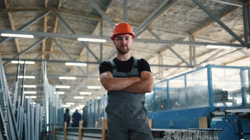 Worker in grey uniform and hard hat stands indoors in the factory with arms crossed. Worker in grey uniform and hard hat stands indoors in the factory with arms crossed.