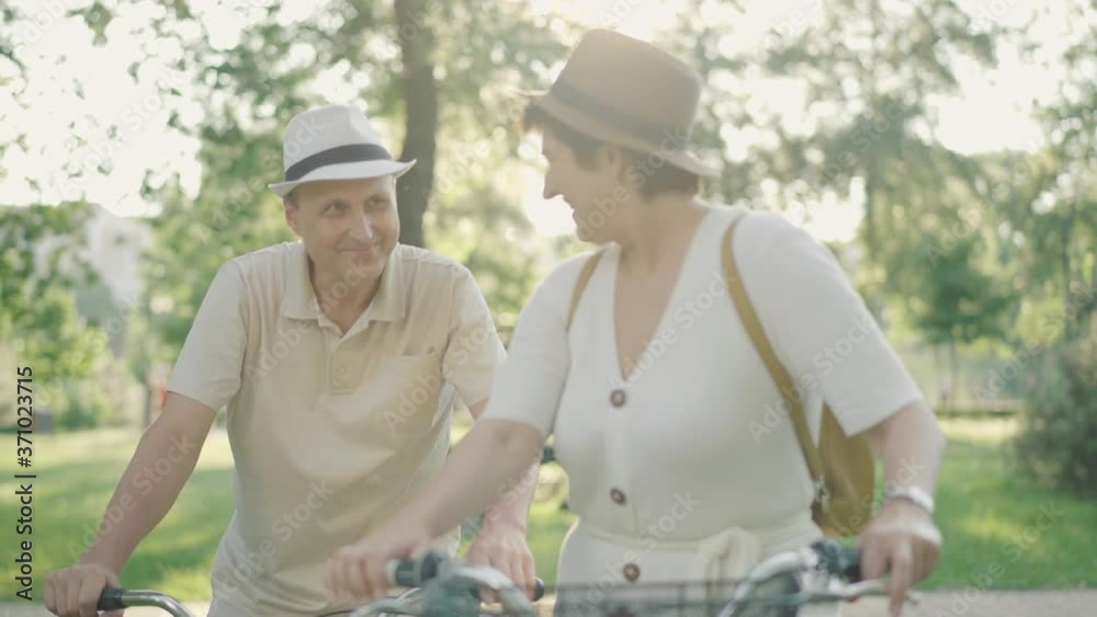 Rack focus changes from charming smiling mid-adult woman looking back to happy loving man riding to her on bike. Cheerful Caucasian couple having fun dating outdoors at sunset. Rack focus changes from charming smiling mid-adult woman looking back to happy loving man riding to her on bike. Cheerful Caucasian couple having fun dating outdoors at sunset.