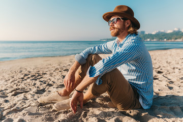 young attractive hipster man sitting on beach by sea on summer vacation, boho style outfit, dressed in shirt, sunglasses and hat