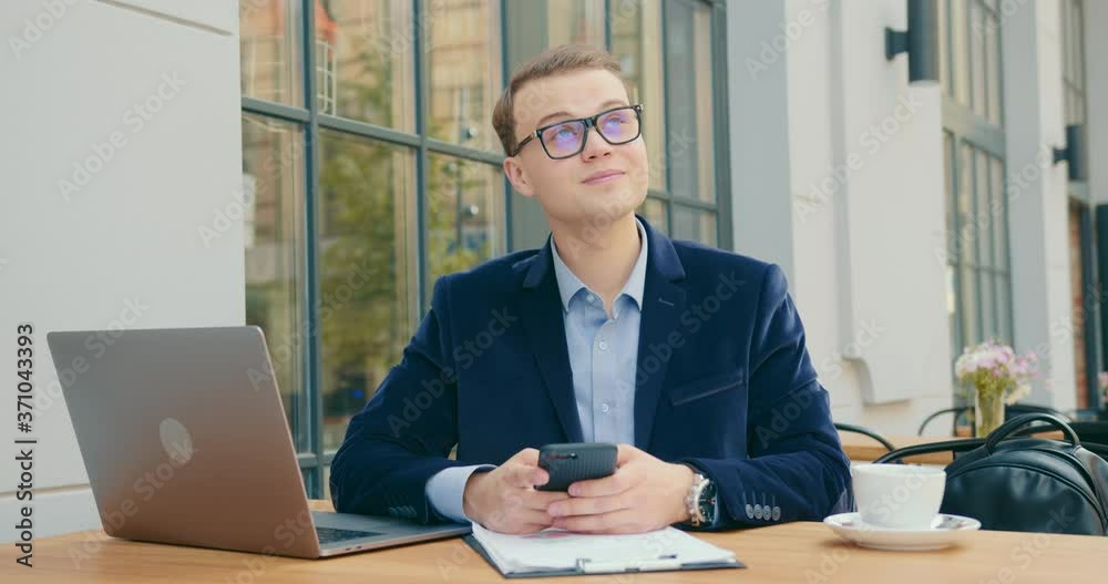 A businessman is sitting at a table in a cafe and texting on his smartphone. He is looking up and smiling. On the table is a laptop and working documents. 4K