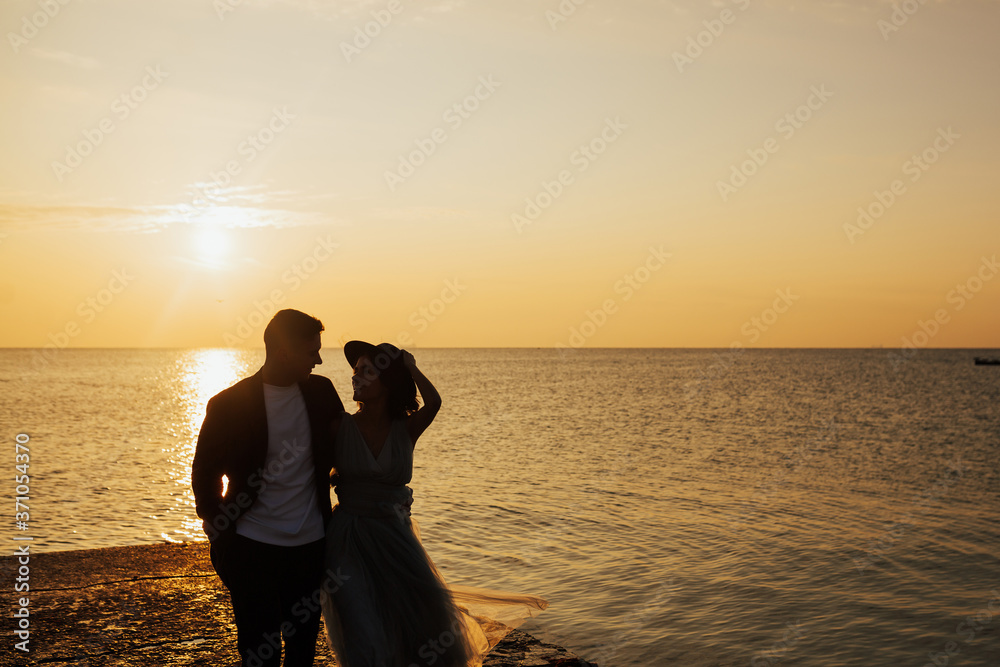 Elegant gorgeous stylish bride with black hat and groom walking in sunset on the background of the sea. Bride and groom walking to the evening wedding ceremony near the sea.
