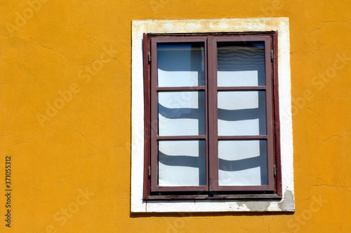 painted brown wooden window in vertical orientation in yellow color stucco exterior wall. bright summer light. European traditional facade detail in old town. residential home. shadows and lights. 